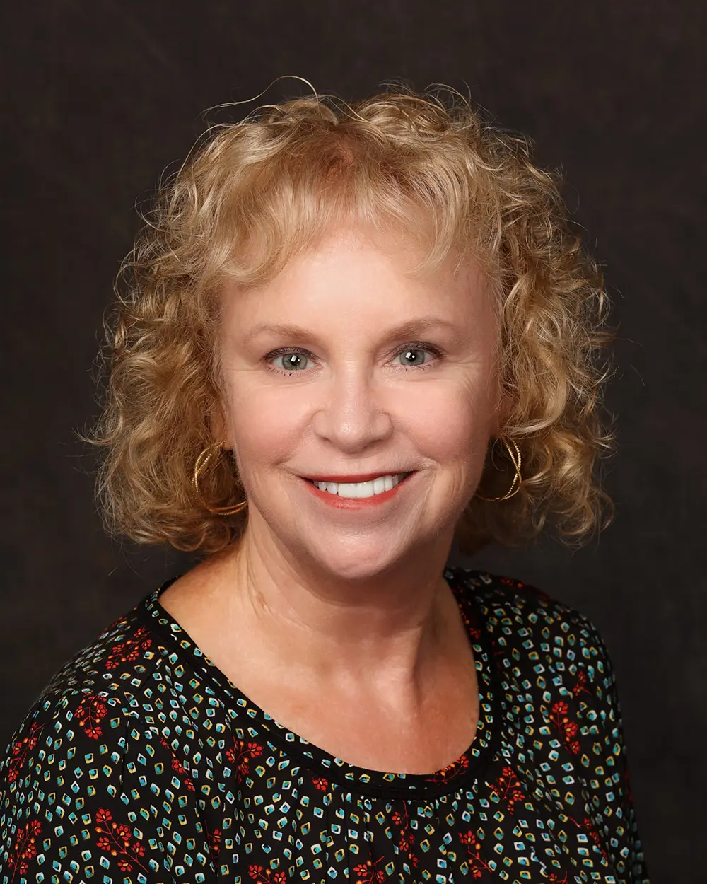 Woman with blond, curly hair smiles into camera in front of dark background