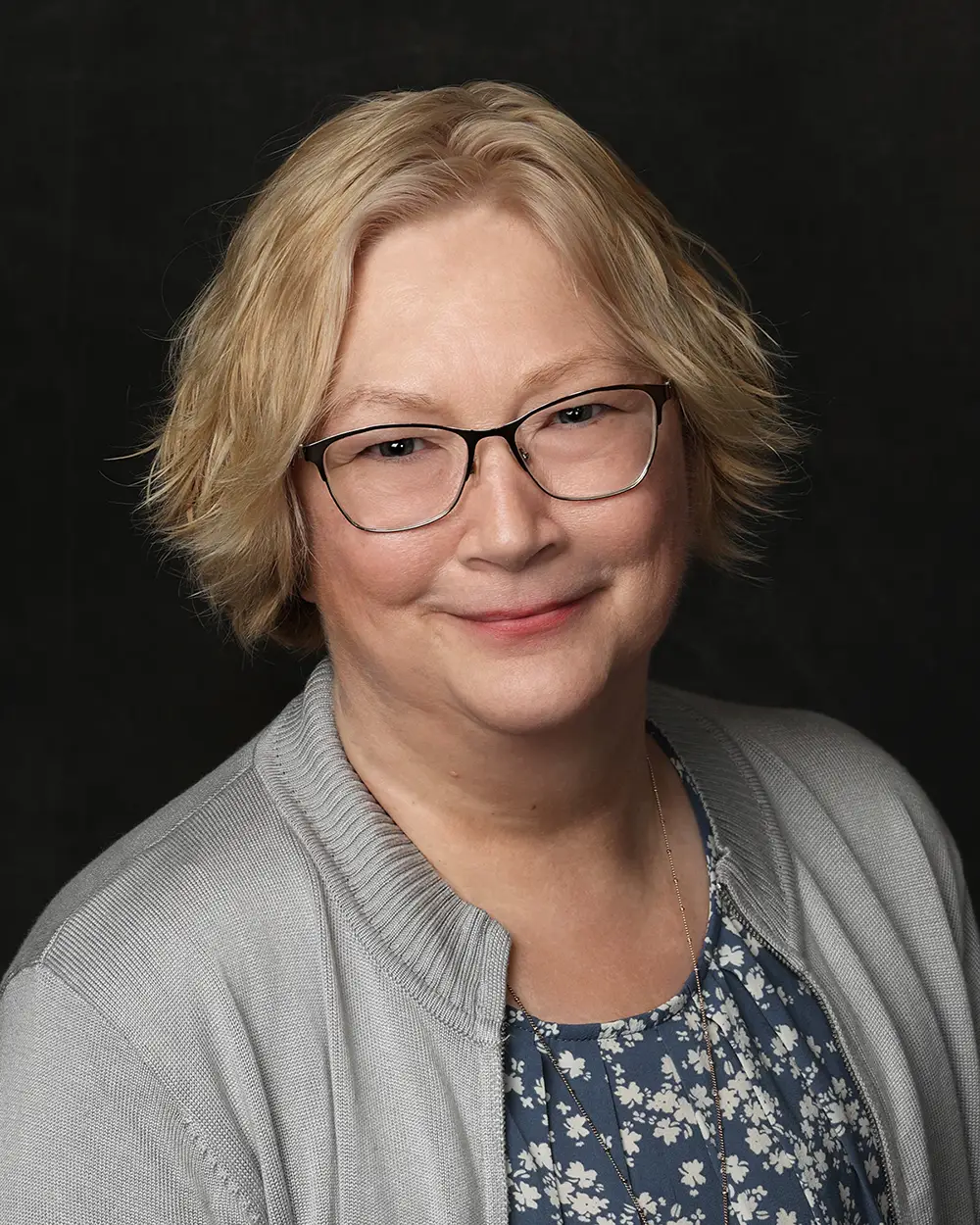 Woman with blond hair, wearing classes, smiling slightly into the camera. Black background.