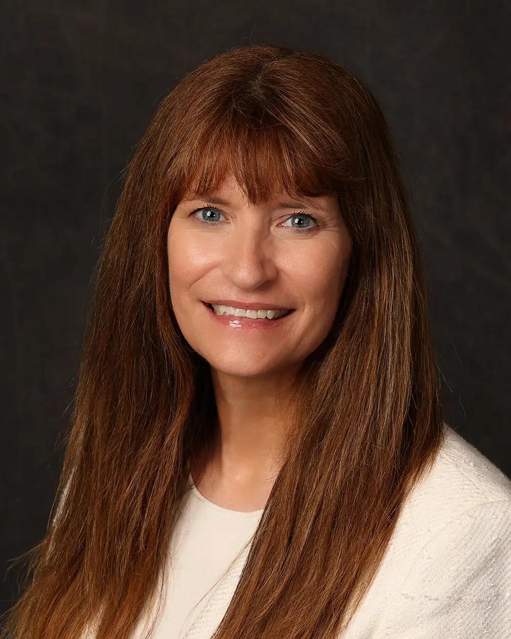 Woman with long, straight, brown hair smiling into camera in front of black background