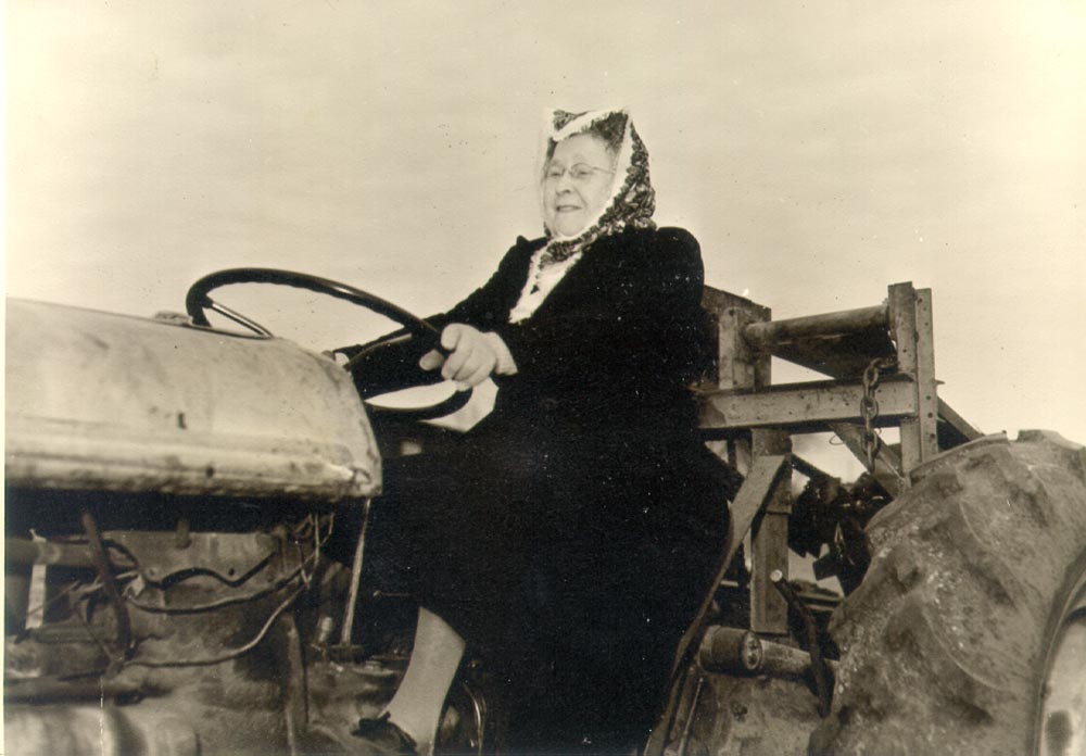 An elderly woman wearing a headscarf is driving a tractor in an open field.
