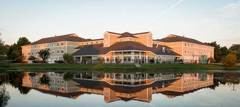 A large building with multiple sections is reflected in a calm body of water, surrounded by greenery and trees under a clear sky. A large building with multiple sections is reflected in a calm body of water, surrounded by greenery and trees under a clear sky.