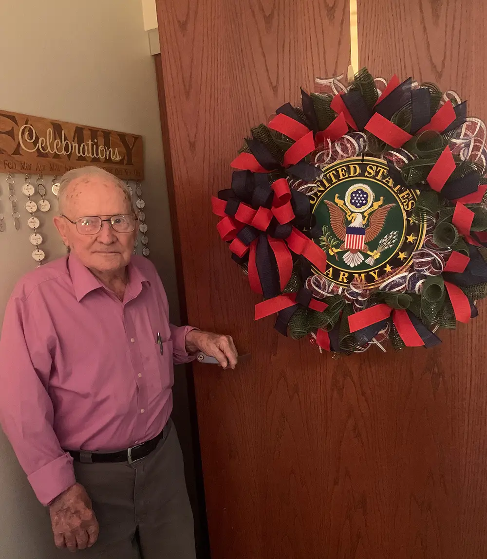 Elderly man standing next to a door decorated with a large patriotic wreath featuring the U.S. Army emblem.