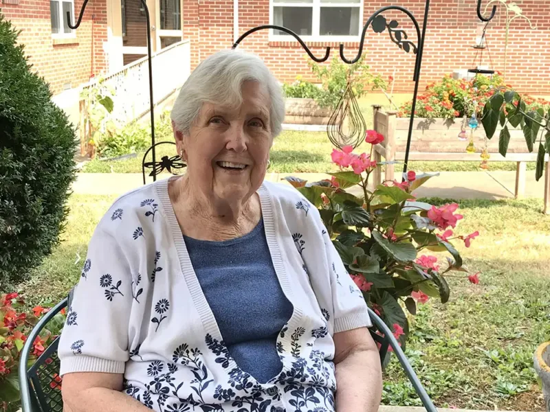 Elderly woman with white hair smiles at camera while sitting in front of potted plants outside
