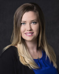 A woman with long, straight hair smiles at the camera, wearing a blue top and a black jacket against a dark background.