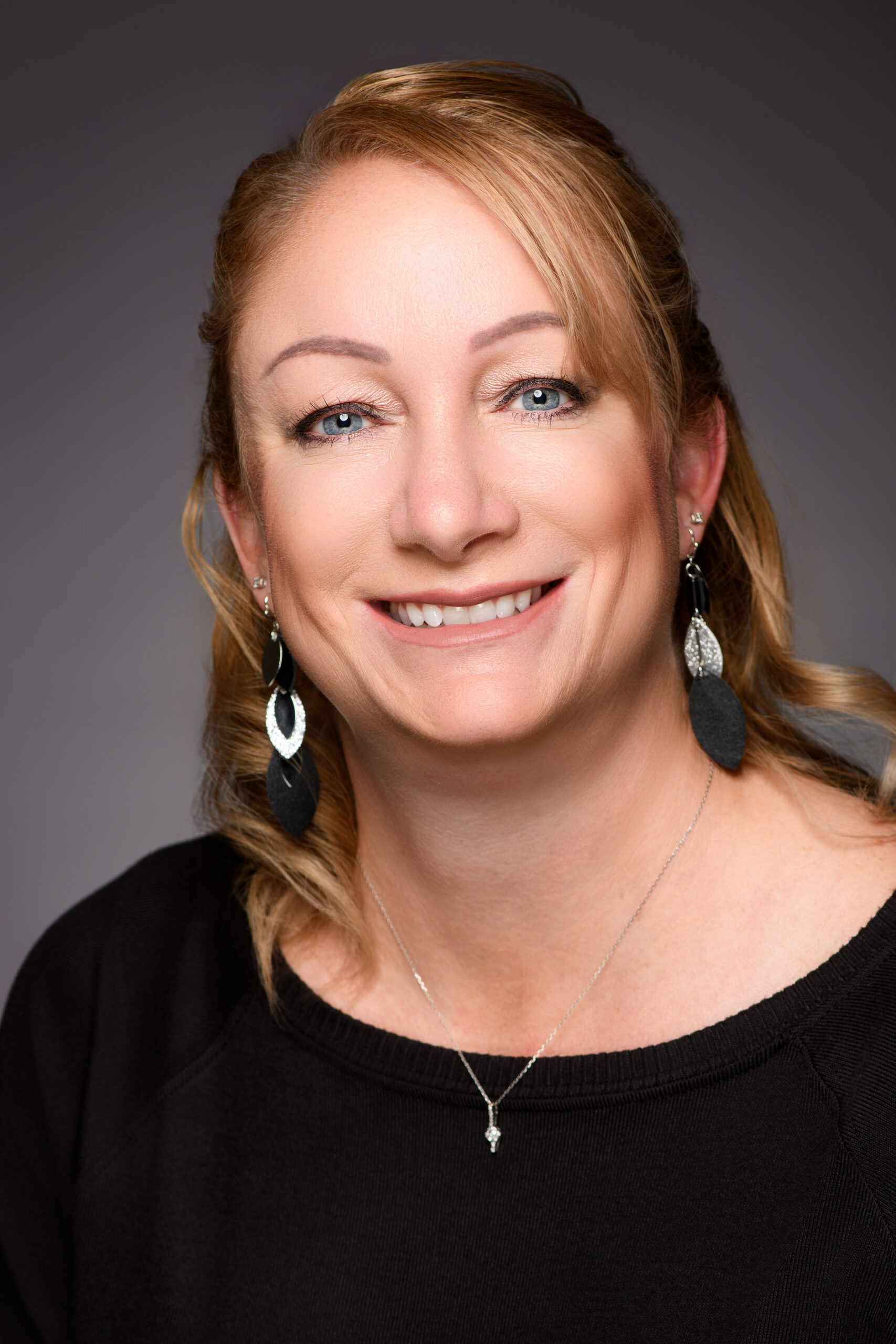 Alisha Craft "Smiling woman with blue eyes and light brown hair wearing black top, silver earrings, and necklace on neutral background