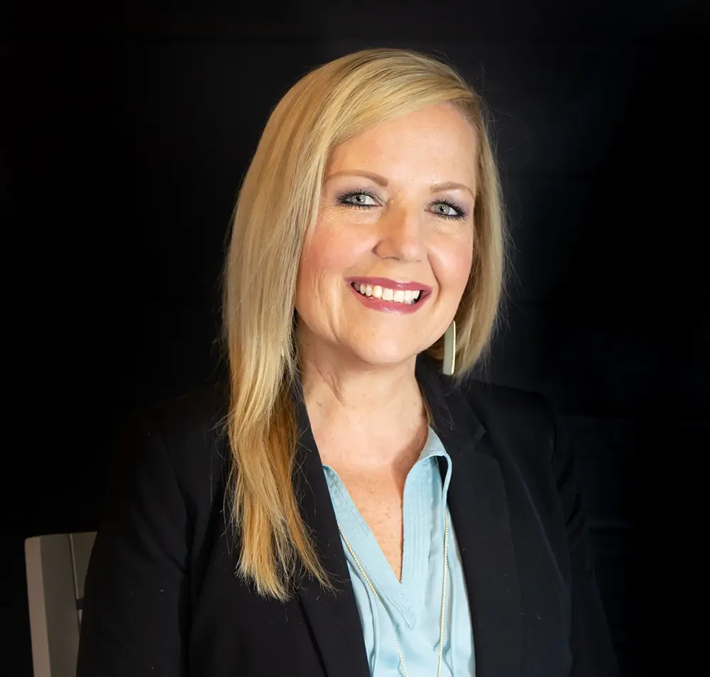 Woman with straight blonde hair smiles into the camera in front of a black background. She's dress professionally -- blouse and blazer