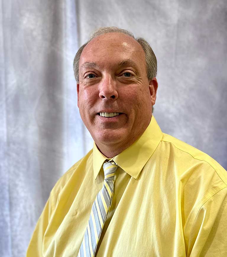 A man with short gray hair, wearing a yellow shirt and a striped tie, smiles at the camera in front of a gray backdrop. A man with short gray hair, wearing a yellow shirt and a striped tie, smiles at the camera in front of a gray backdrop.