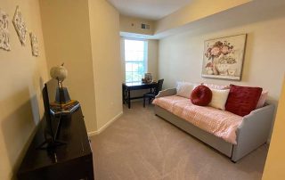 A small, cozy room with a gray daybed, pink bedding, two red pillows, a black desk with a chair, a painting of flowers above the daybed, and beige carpeted flooring. A small table with a globe is in the foreground.
