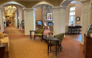 A hotel lobby with white archways, green and brown chairs, small tables, and warm lighting. The lobby extends into a larger area with additional seating. A clock and lamp are visible.