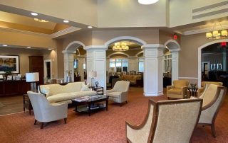 A spacious, elegant lobby with beige and white furniture, modern lighting, and high ceilings. The room features multiple seating areas with cushioned chairs and sofas on a red-patterned carpet.