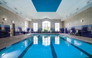 Indoor swimming pool in a facility with rows of lounge chairs and potted plants along the sides, under a high ceiling. Large windows at the far end allow natural light to enter.
