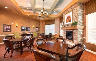 A cozy room with multiple tables and leather chairs, a stone fireplace, framed pictures on the walls, and ceiling lights.