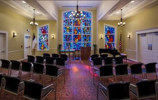 A small chapel with rows of chairs facing a podium, a piano to the side, and colorful stained glass windows at the front.
